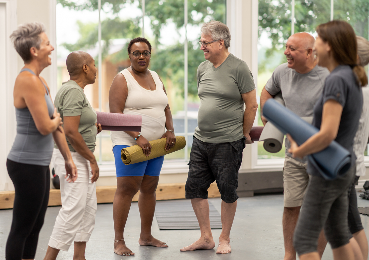 A diverse group of adults stand around after class with their yoga mats in their arms and socialize.